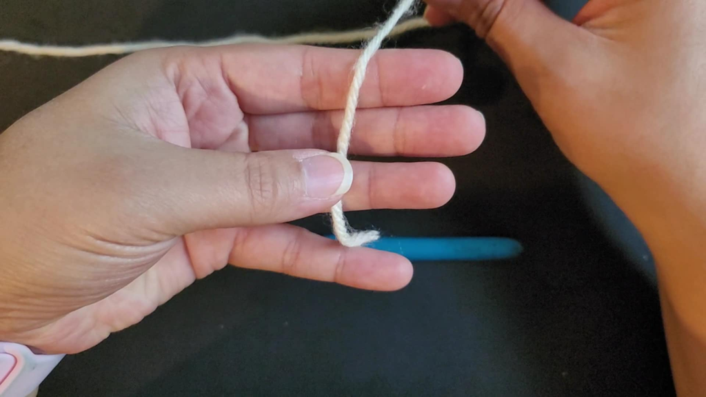 picture of hands holding a single strand of yarn with the yarn secured by the thumb against the ring finger to start the process of making a magic circle