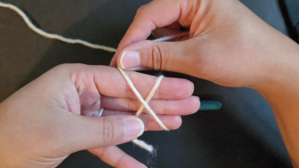 picture of hands holding a single strand of yarn with the yarn secured by the thumb against the ring finger and the yarn wrapped around fingers twice creating an X in front and parallel lines on the back of the hand for a magic circle
