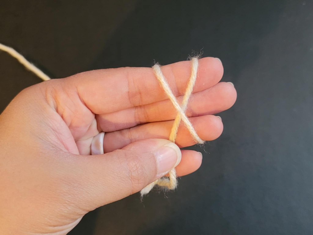 picture of hands holding a single strand of yarn with the yarn secured by the thumb against the ring finger and the yarn wrapped around fingers twice creating an X in front and parallel lines on the back of the hand for a magic circle