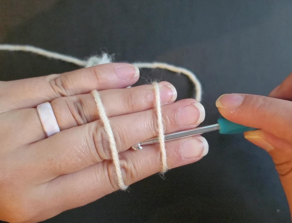 picture of back of hand with 2 parallel yarn strands showing and the crochet hook going under the first strand closest to the finger tips.